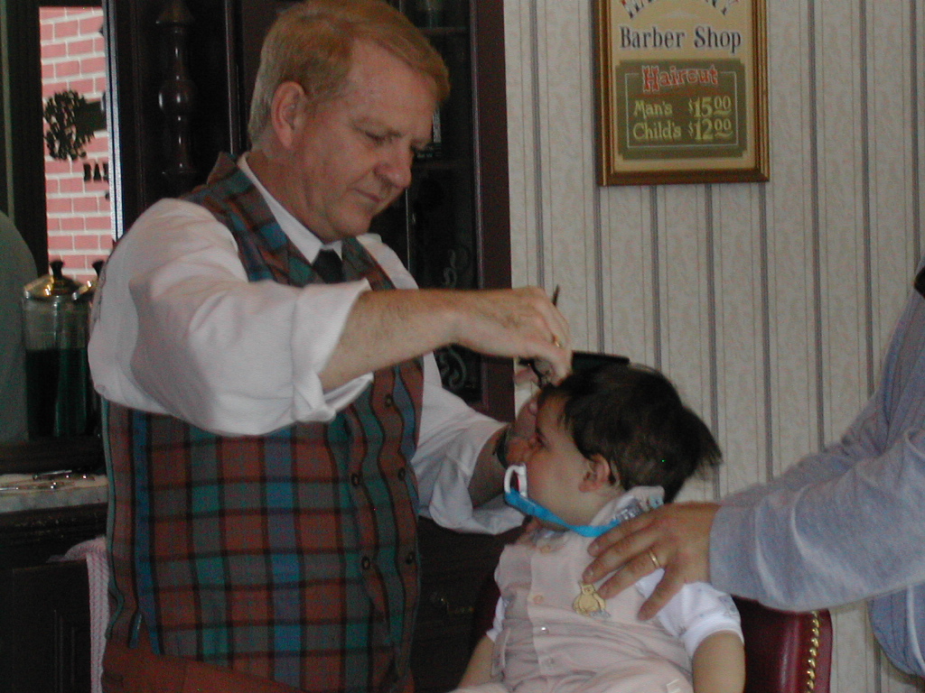 a small child getting a haircut in the magic kingdom. The barber is a blondish man wearing a red and green plaid vest over a white button down shirt. The child is wearing tan overalls over a white shirt. The child has a pacifier in his mouth.